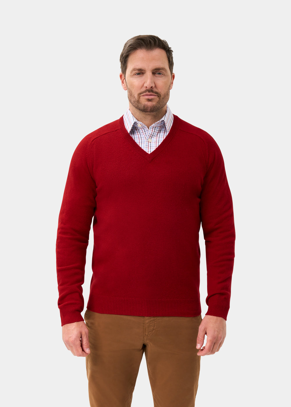 Man wears Hampshire Lambswool Vee Neck Jumper in Dubonnet over plaid shirt, brown pants; plain light background.