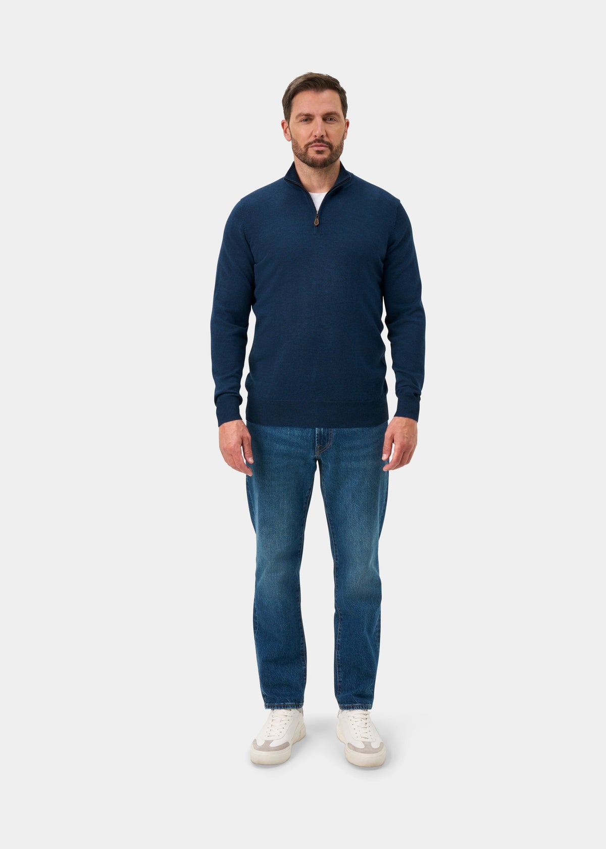 Man in a Barton Merino Wool Quarter Zip Jumper in Indigo, blue jeans, white sneakers; plain light background.