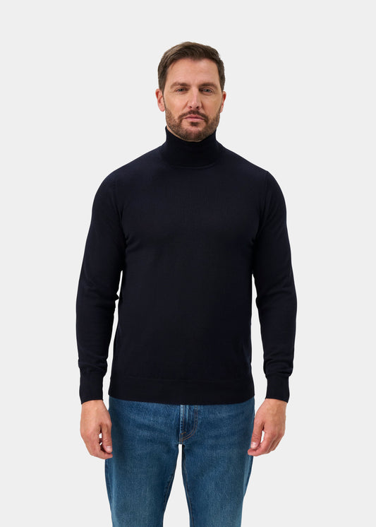 Man wears Linton Merino Wool Roll Neck Jumper in Dark Navy, paired with blue jeans against a plain background.