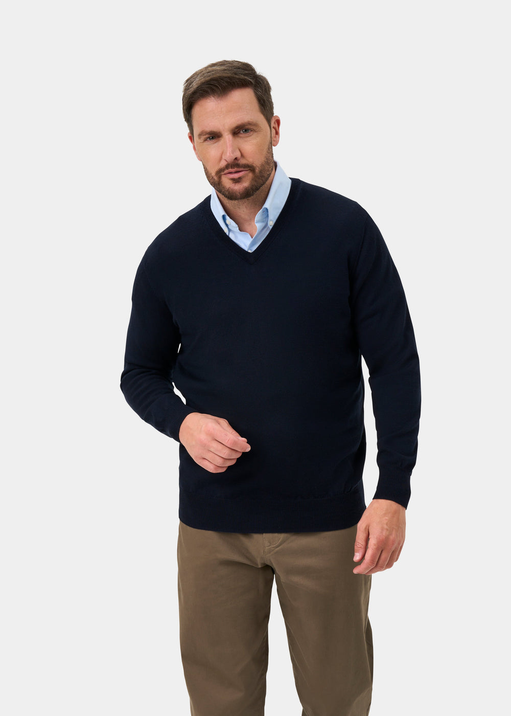 A man wearing a dark navy blue v-neck sweater over a light blue collared shirt and brown trousers, standing against a light gray studio background.