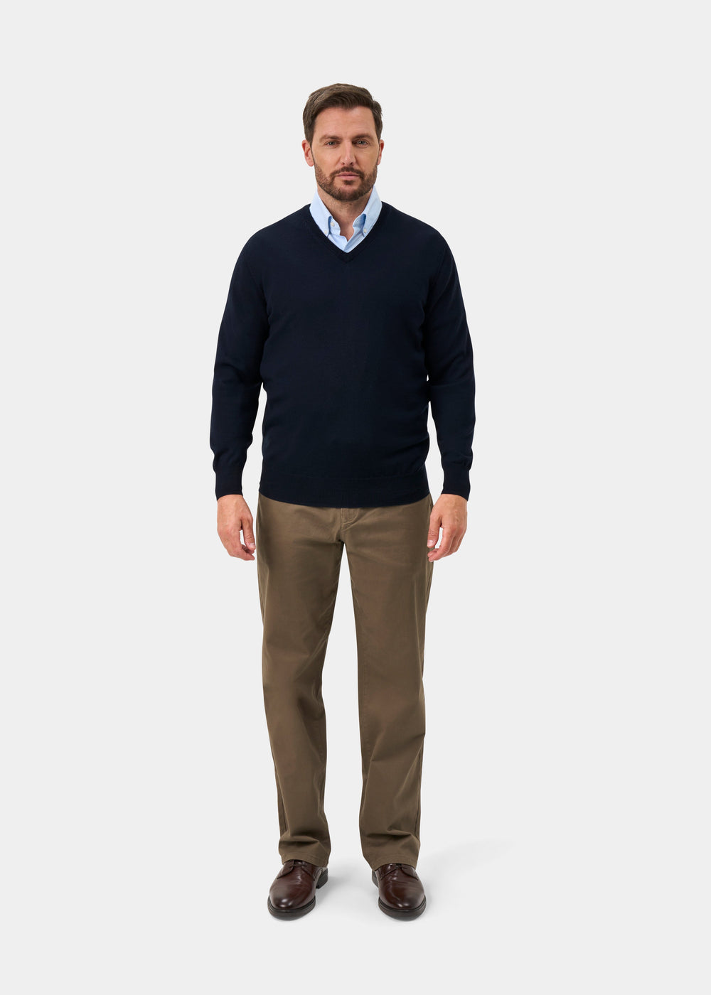 A man wearing a navy blue v-neck sweater over a light blue collared shirt, paired with brown trousers and dark brown leather shoes, standing against a light gray studio background.