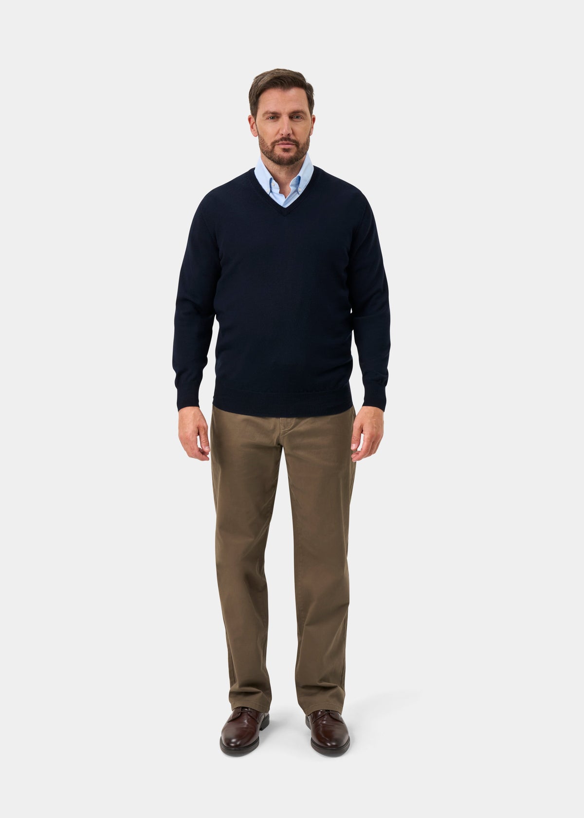 A man wearing a navy blue v-neck sweater over a light blue collared shirt, paired with brown trousers and dark brown leather shoes, standing against a light gray studio background.