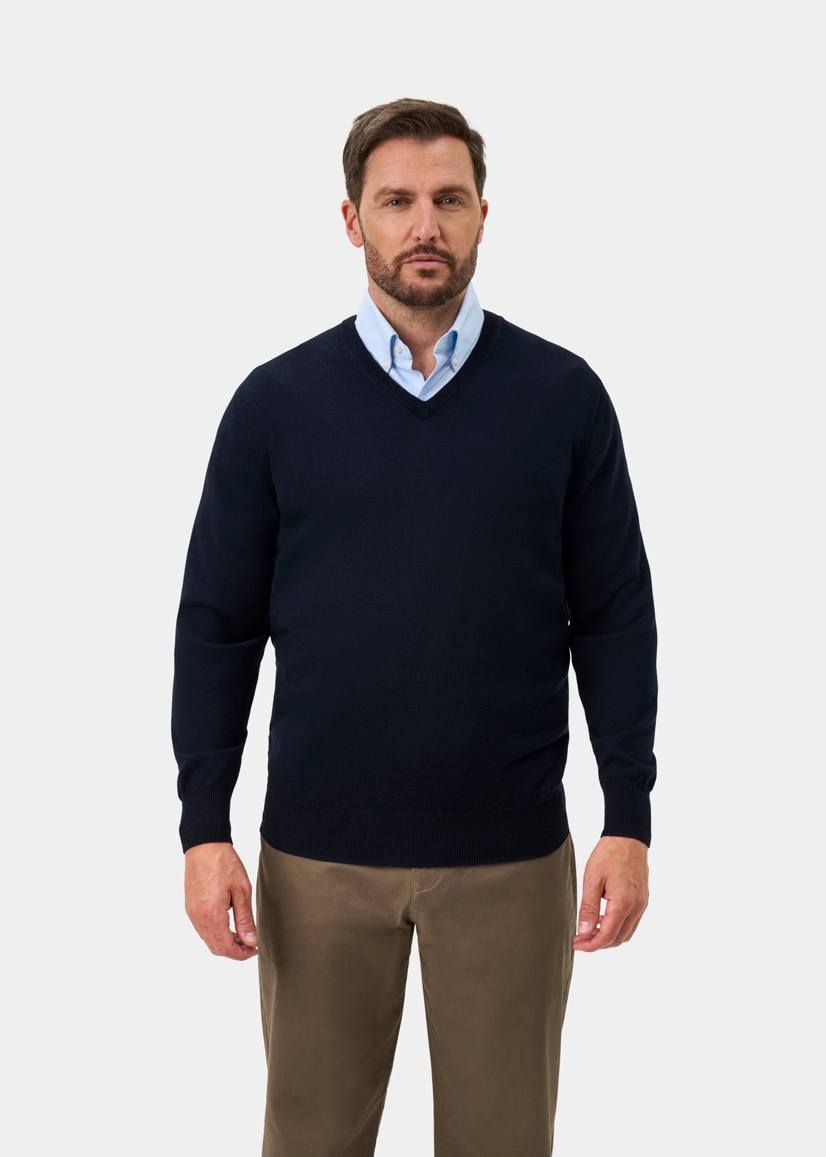 A man wearing a navy blue v-neck sweater over a light blue collared shirt and brown trousers, standing against a light gray studio background.