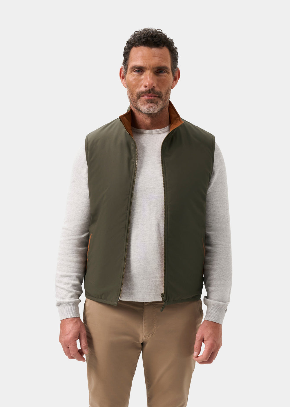 A man wearing a Swifthall Reversible City Gilet In Olive and Cider over a light gray sweater and tan pants.