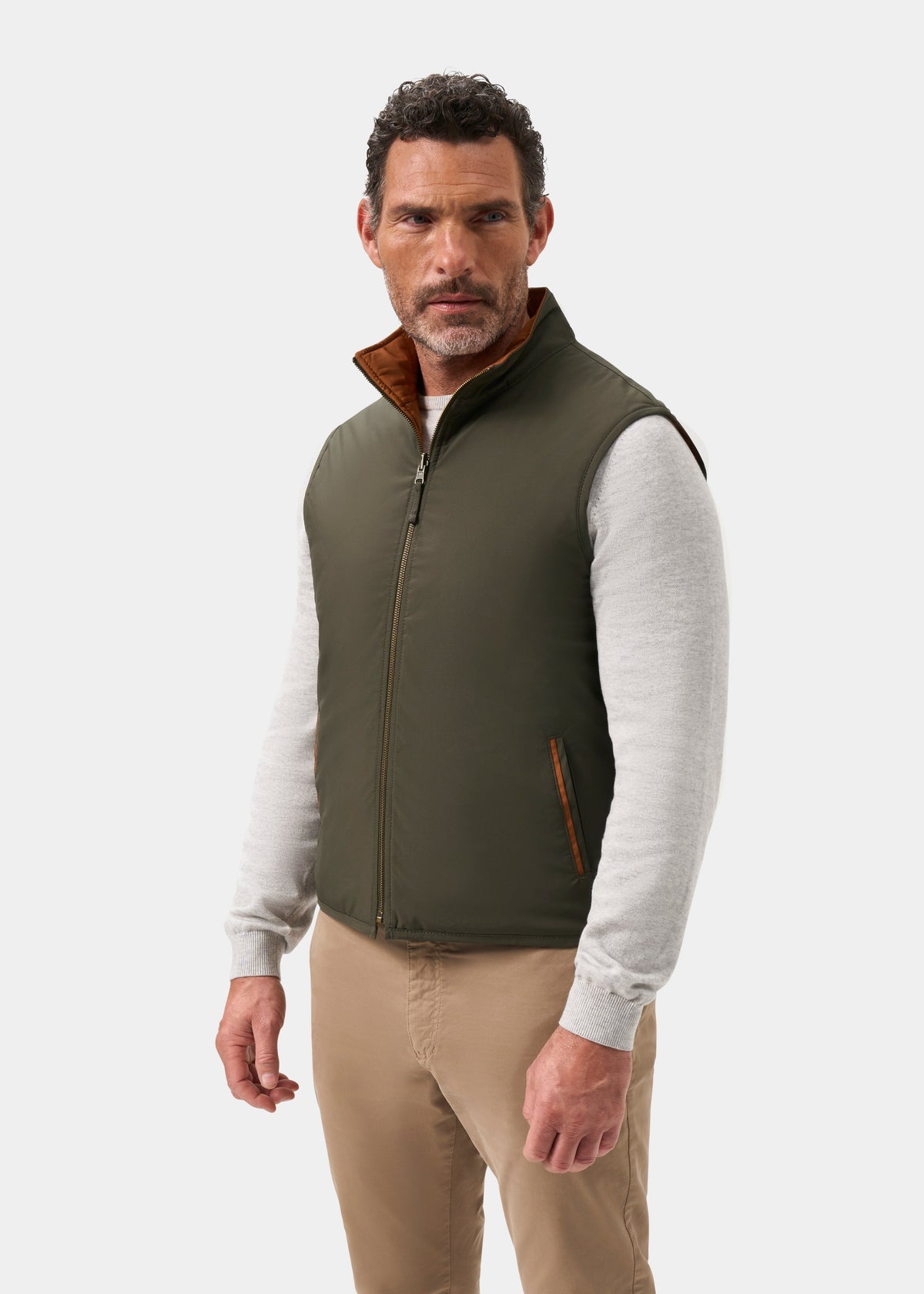 A man wears a Swifthall Reversible City Gilet In Olive and Cider over a light gray sweater and tan trousers.