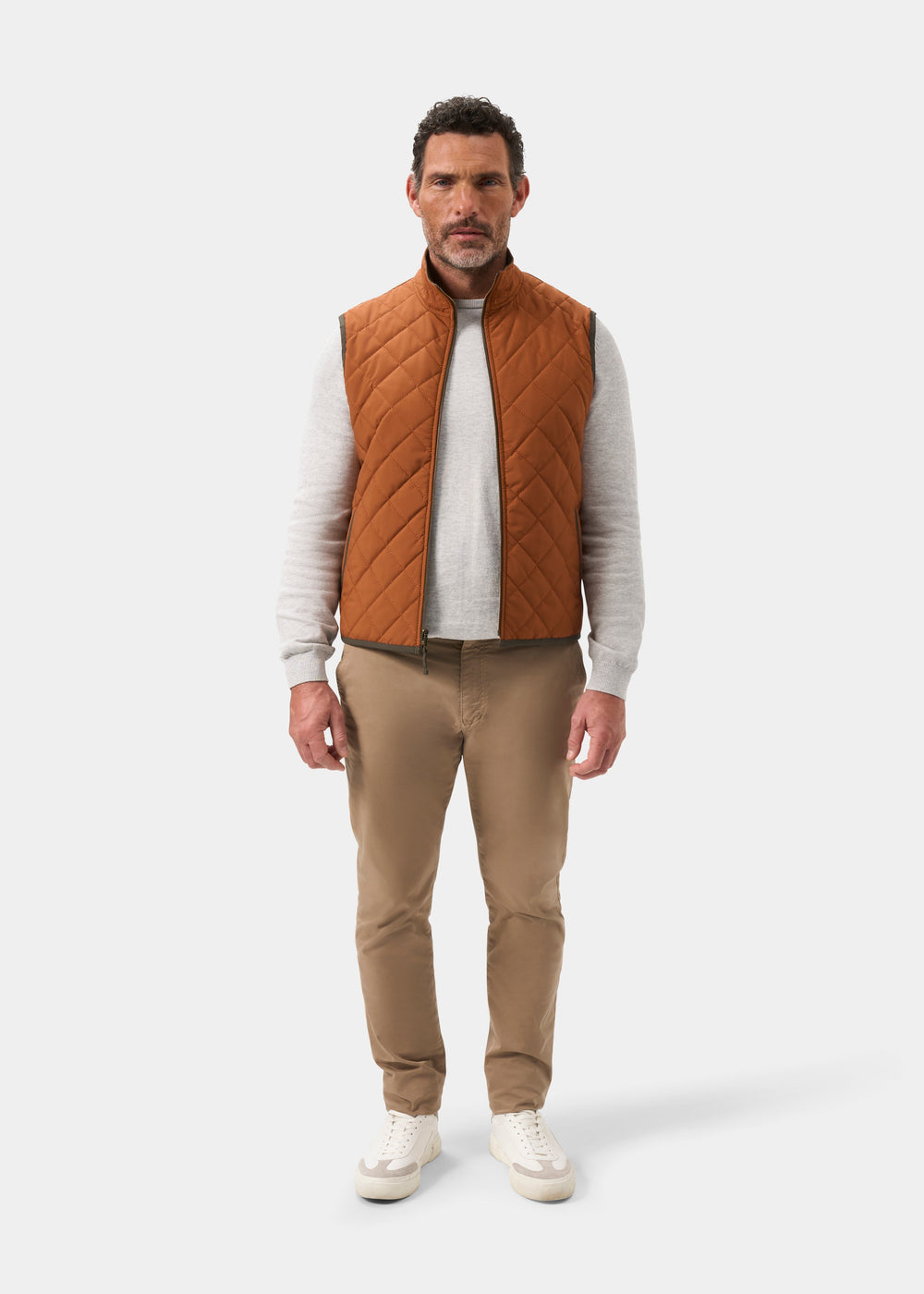 A man wears a Swifthall Reversible City Gilet In Olive and Cider over a gray sweater, tan trousers, and white sneakers.