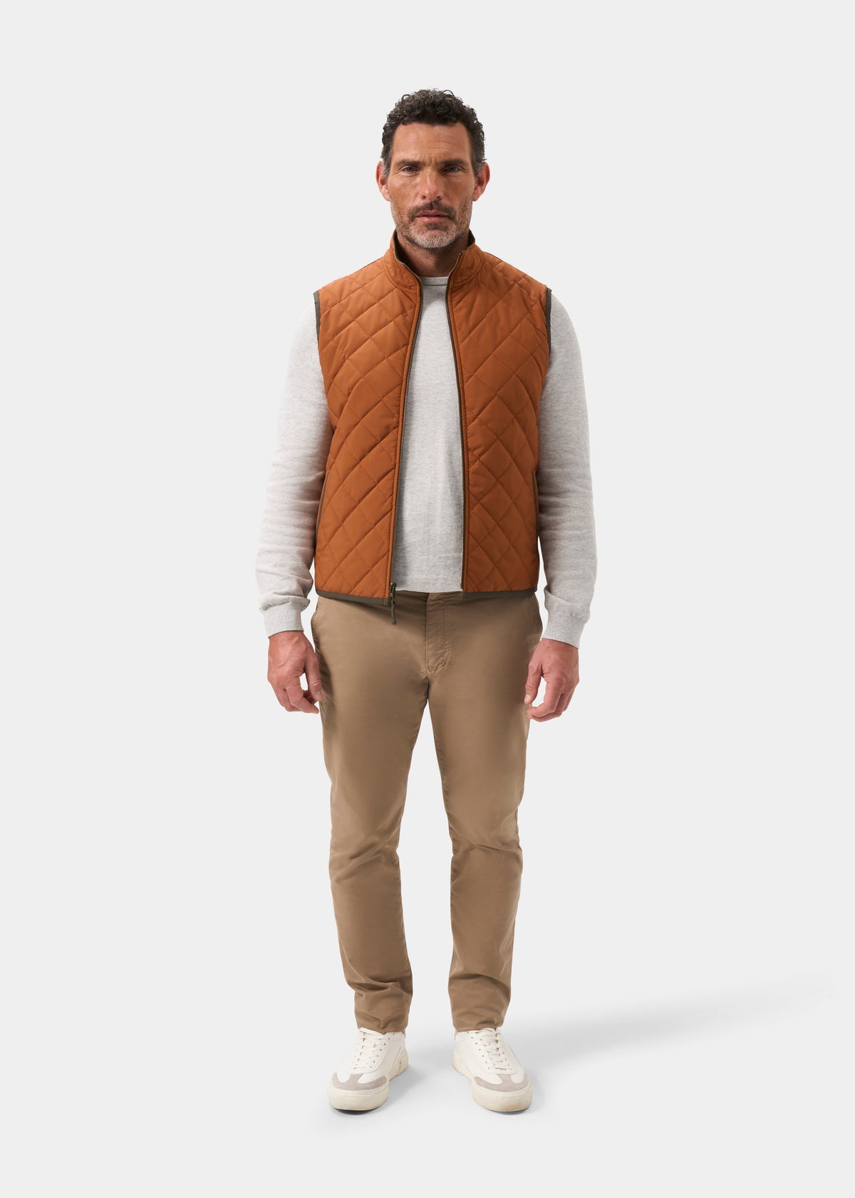 A man wears a Swifthall Reversible City Gilet In Olive and Cider over a gray sweater, tan trousers, and white sneakers.