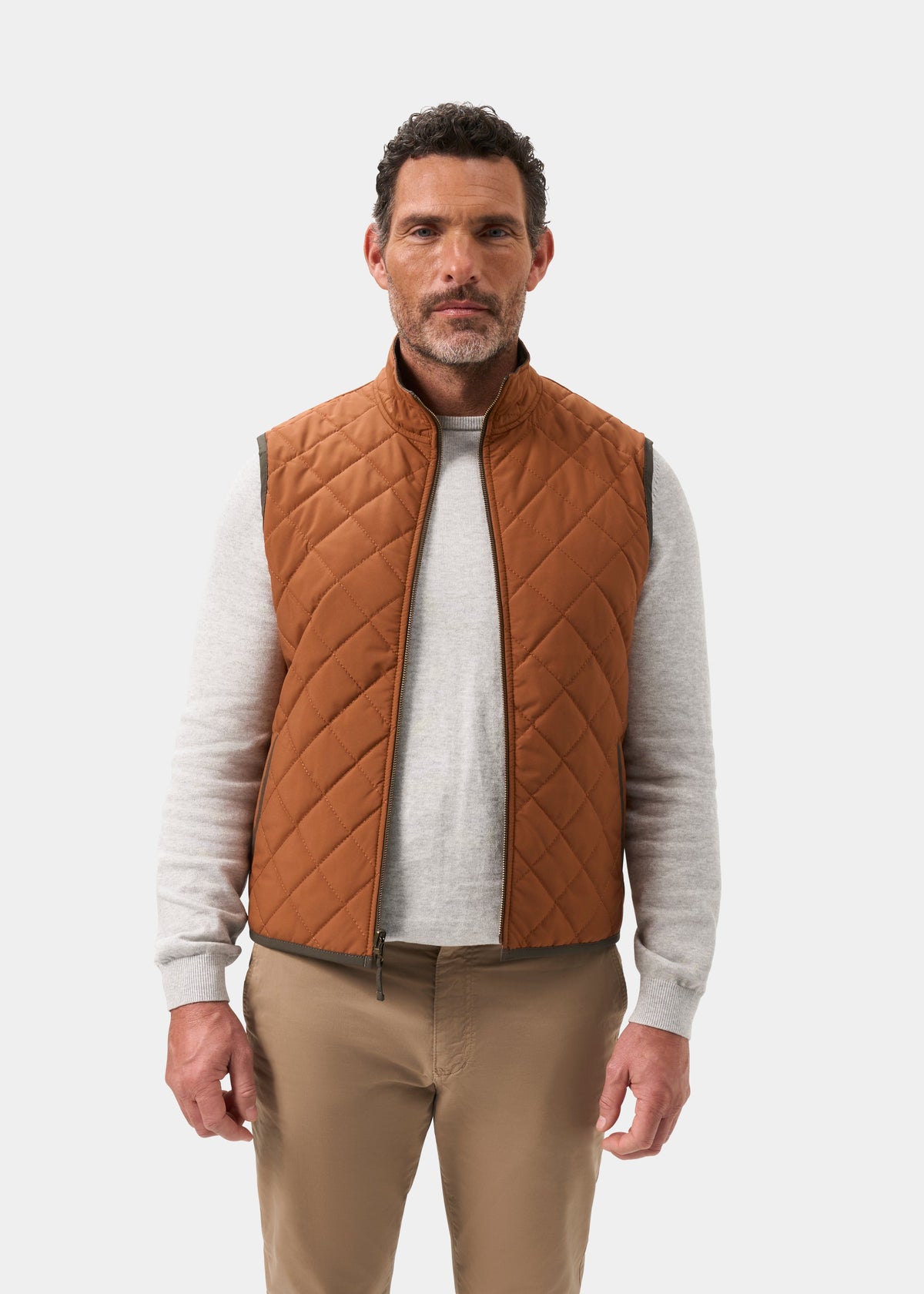 A man wears a cider-colored Swifthall Reversible City Gilet In Olive and Cider over a gray shirt and tan trousers.