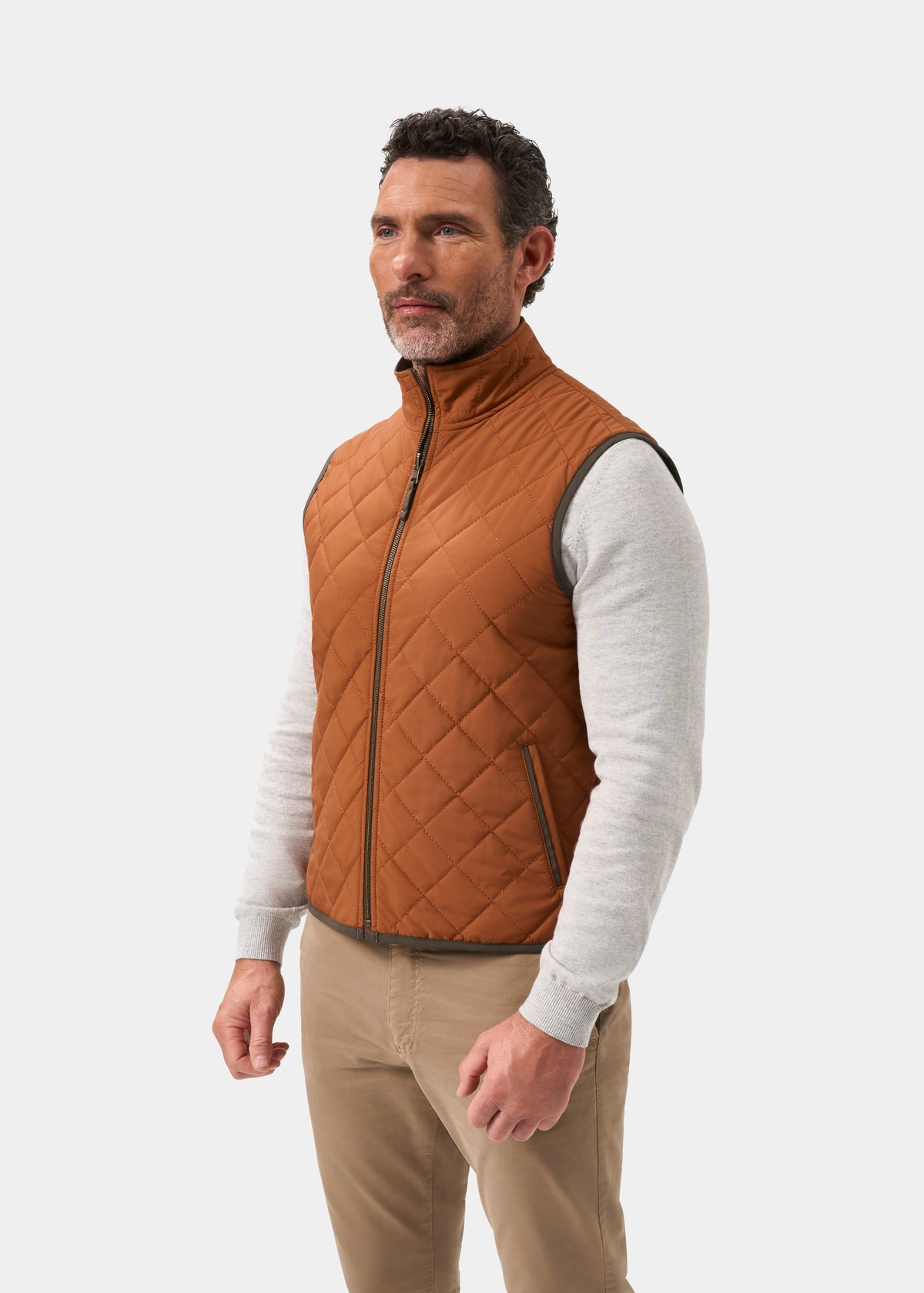 A man wears a cider-colored Swifthall Reversible City Gilet In Olive and Cider over a light gray sweater and tan trousers.