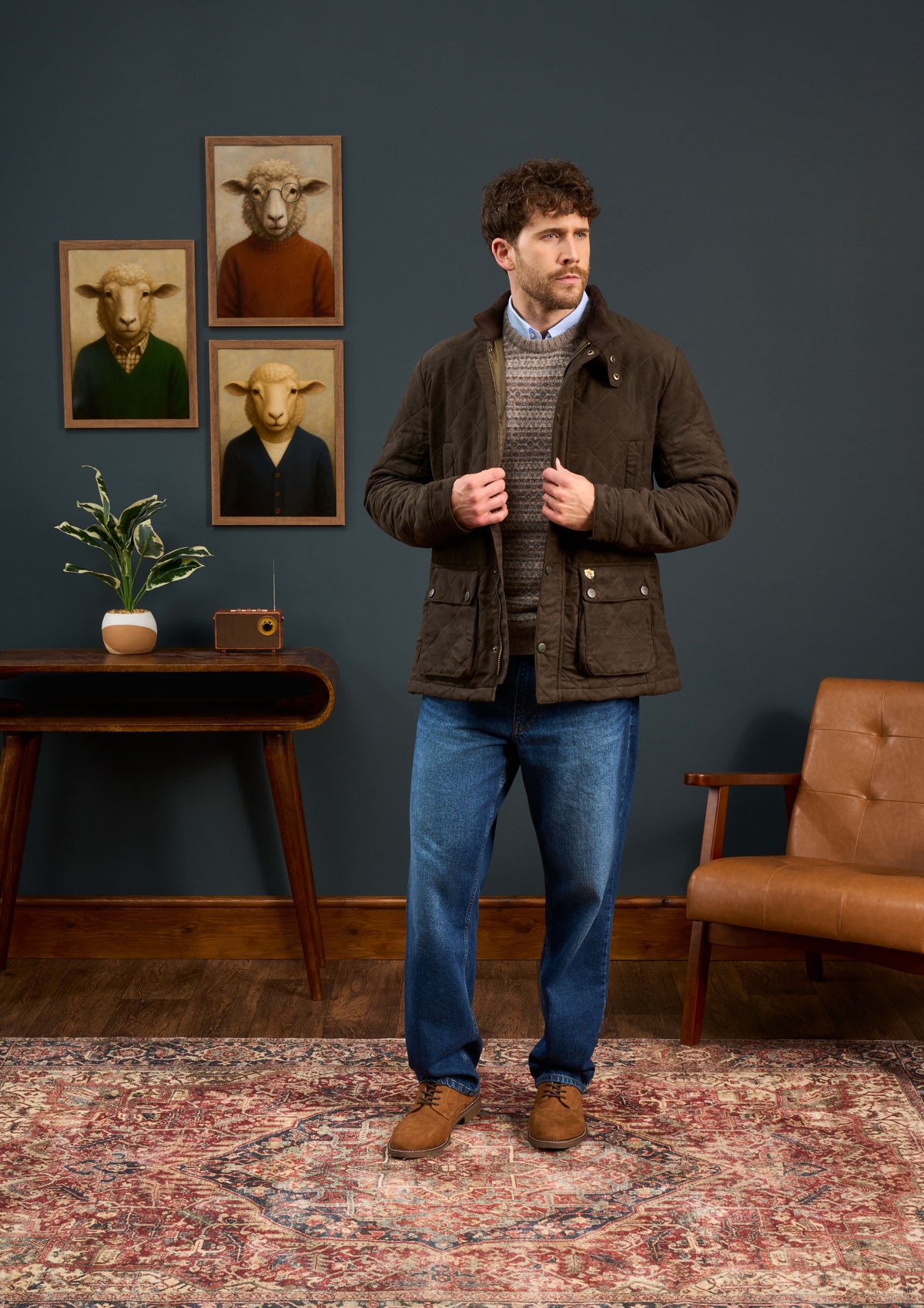 Man wears Felwell Children’s Quilted Jacket in Olive, regular fit, with jeans in room of animal art and vintage furniture.