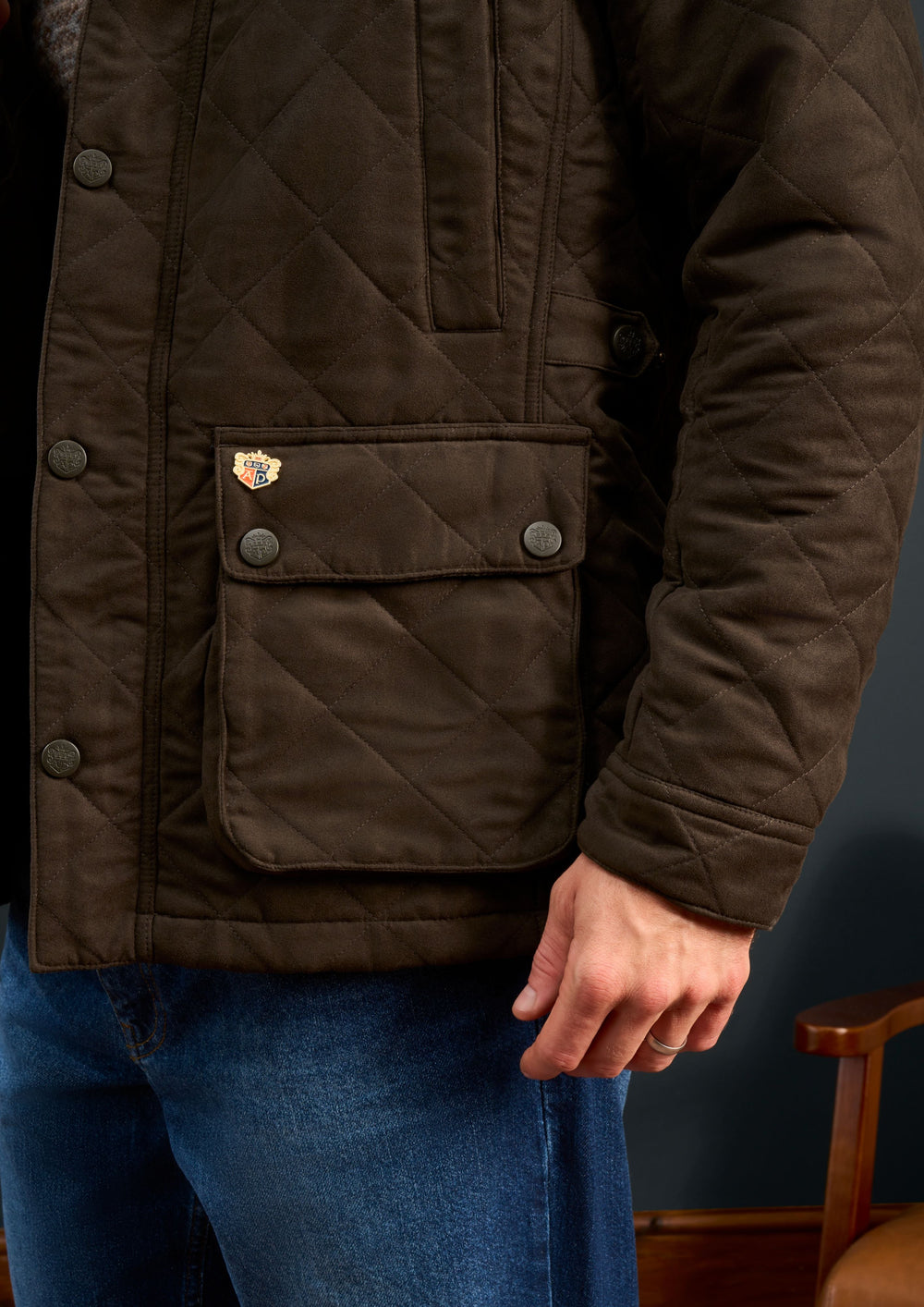 A child in an olive Felwell quilted jacket stands by a wooden chair, hand at their side, wearing blue jeans.