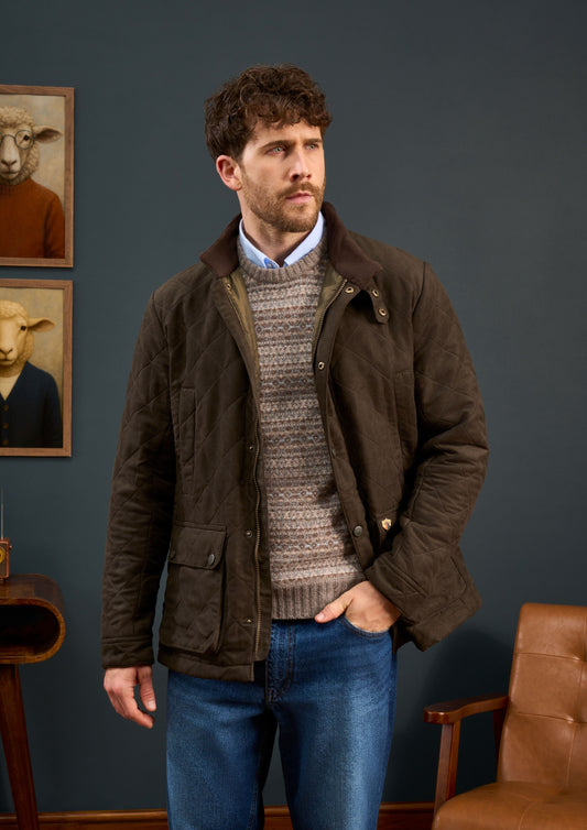 A boy in a Felwell Children’s Quilted Jacket in Olive stands indoors near a chair and animal portraits on a dark wall.