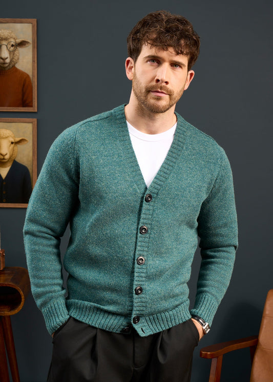 Man in Kinloch Supersoft Shetland Wool V-Neck Cardigan in Kingfisher stands indoors, sheep portraits behind him.