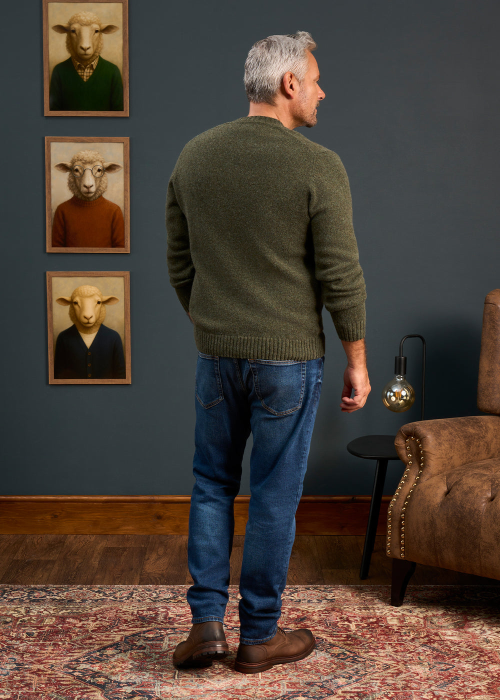 A man in a Kinnadie Supersoft Shetland Wool Crew Neck Jumper stands by a brown chair and three sheep portraits.