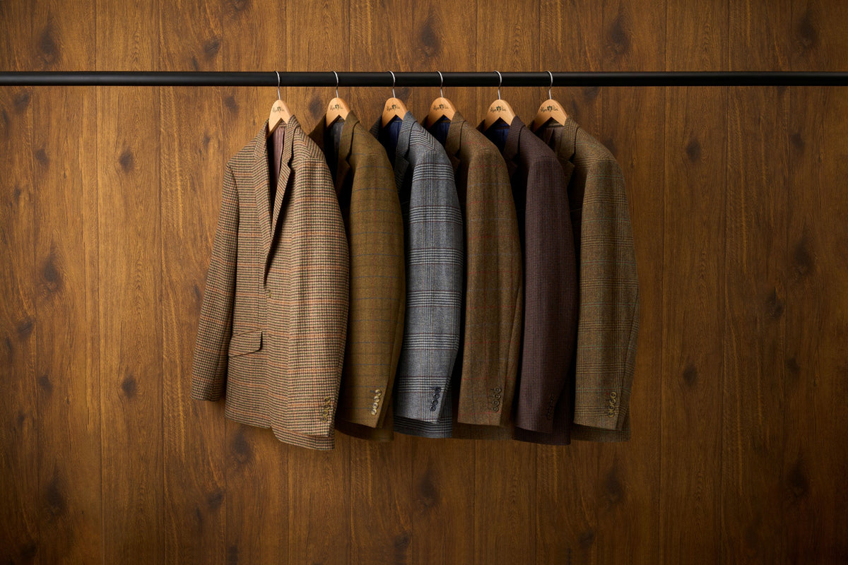 Six men's tweed blazers in various earthy colors (brown, beige, blue, gray, burgundy) hanging on wooden hangers from a black clothing rack against a dark wood-paneled wall.