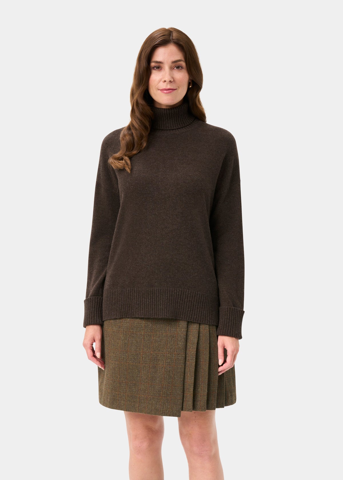 Woman wears Alexa Wool & Cashmere Roll Neck Jumper in Walnut, paired with a pleated skirt, facing forward.