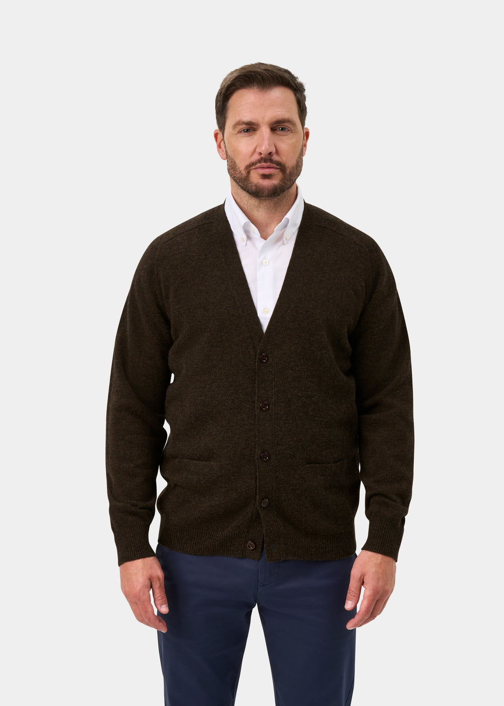 Man in Cornwall Lambswool V-Neck Cardigan in Cocoa, white shirt, navy pants, standing against a plain background.