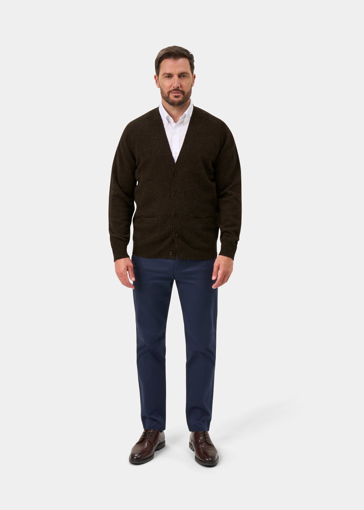 Man in Cornwall Lambswool V-Neck Cardigan in Cocoa, white shirt, blue pants, brown shoes on plain light background.