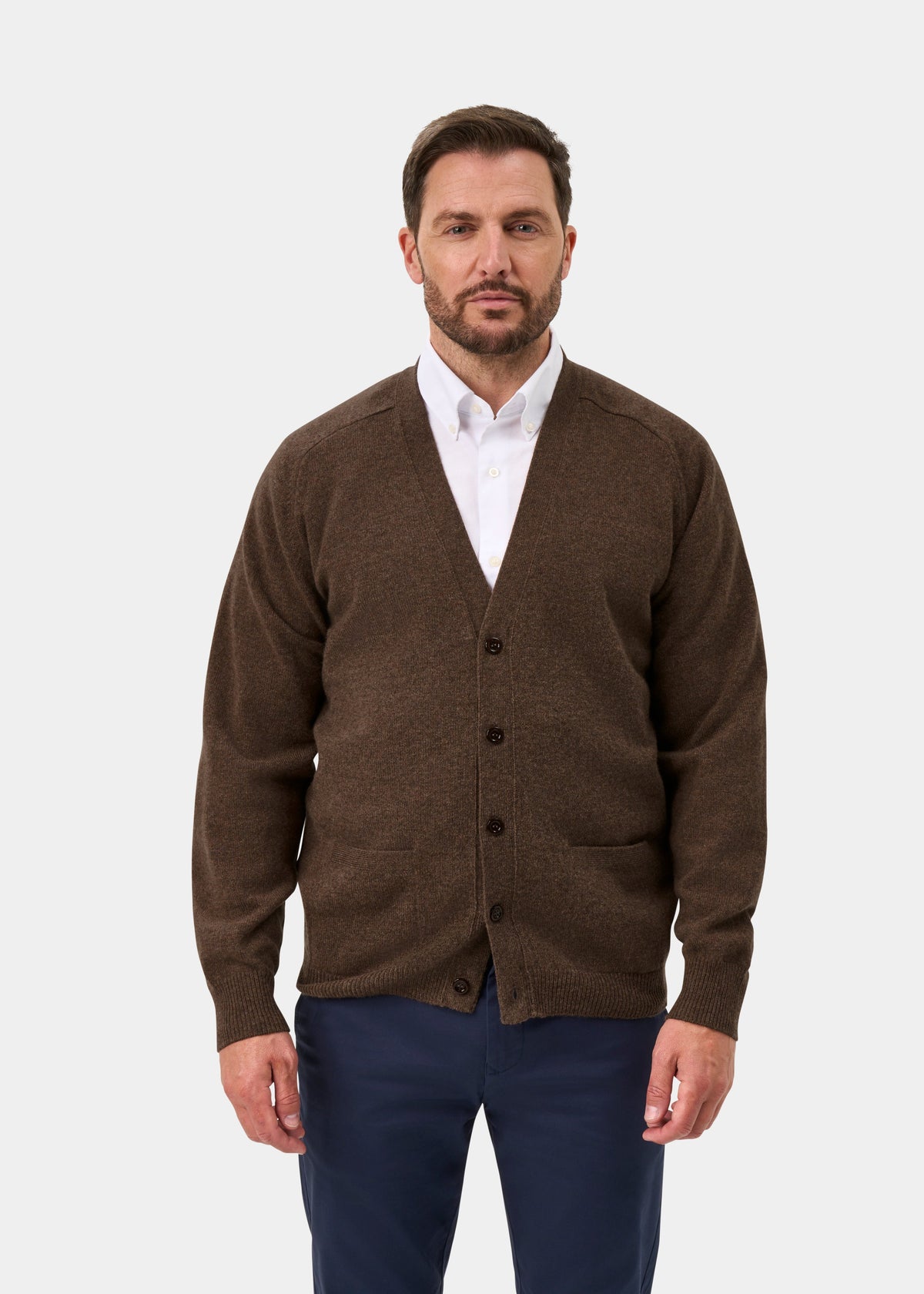 Man in Cornwall Lambswool V-Neck Cardigan in Tobacco, white shirt, navy pants stands facing forward, plain background.