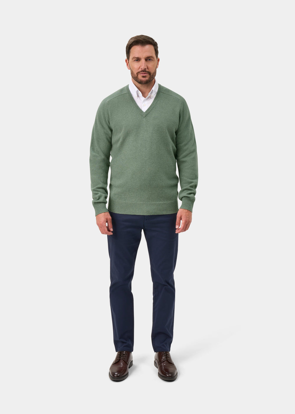 Man in Hampshire Lambswool V-Neck Jumper in Mojito, white shirt, navy pants, and brown shoes against a plain background.