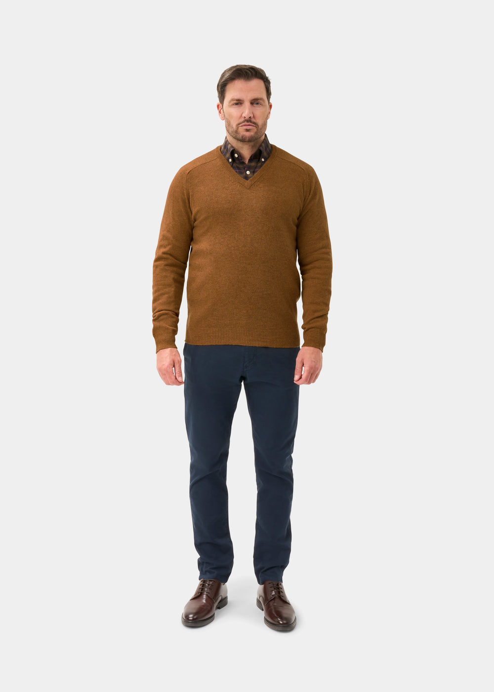 Man in Kilsyth Lambswool V-Neck Jumper in Gazelle, dark shirt, navy pants, and brown shoes on light background.