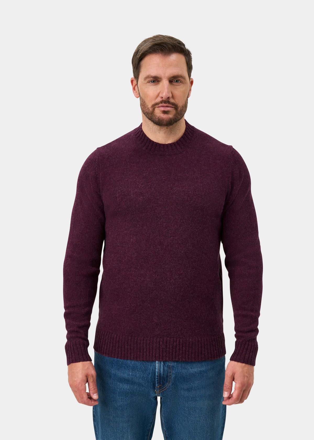 Man wearing a Kinnadie Supersoft Shetland Wool Crew Neck Jumper in Elderberry and blue jeans on white background.