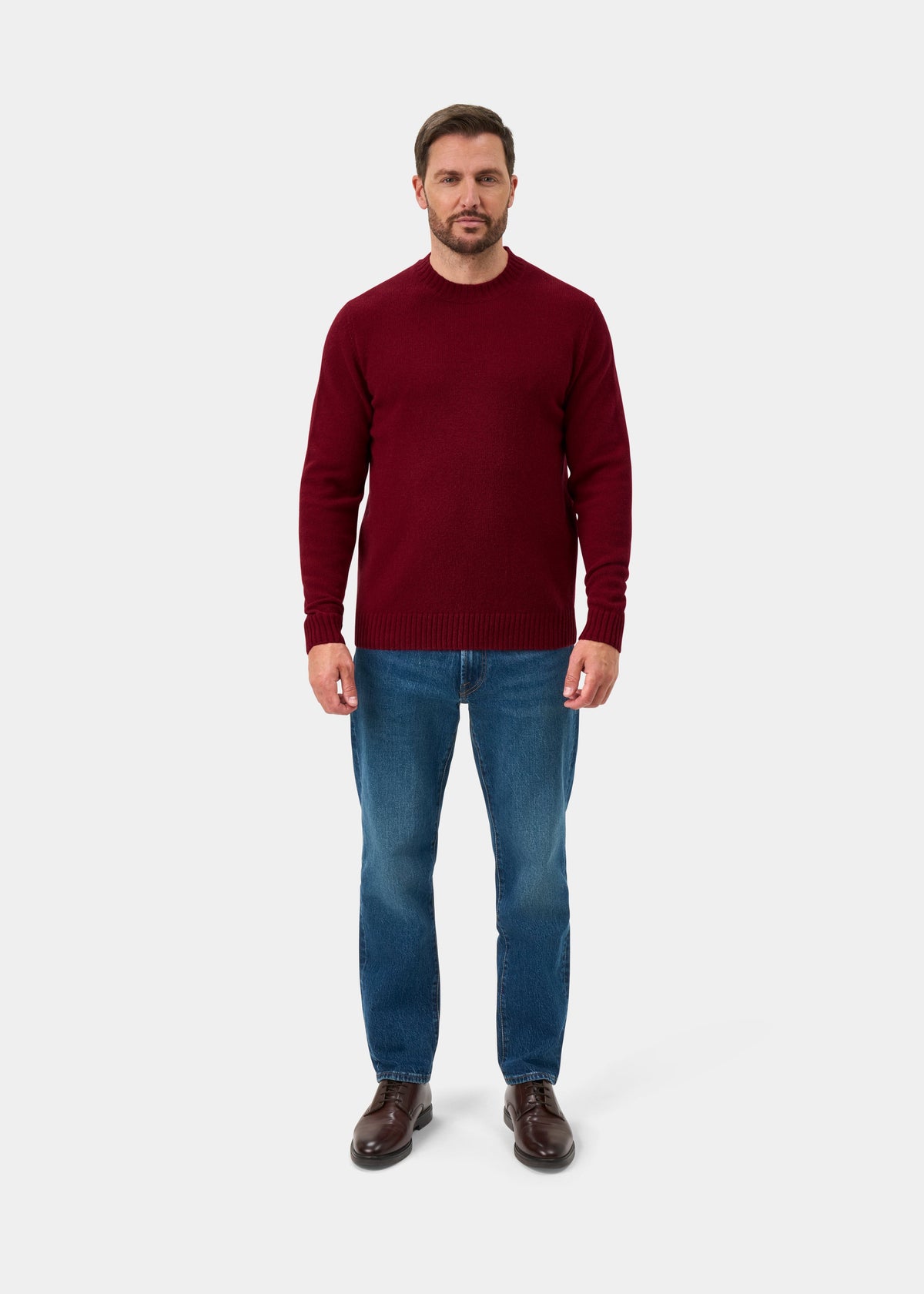 Man wears Kinnadie Supersoft Shetland Wool Crew Neck Jumper in Venetian, blue jeans, and brown shoes on white backdrop.