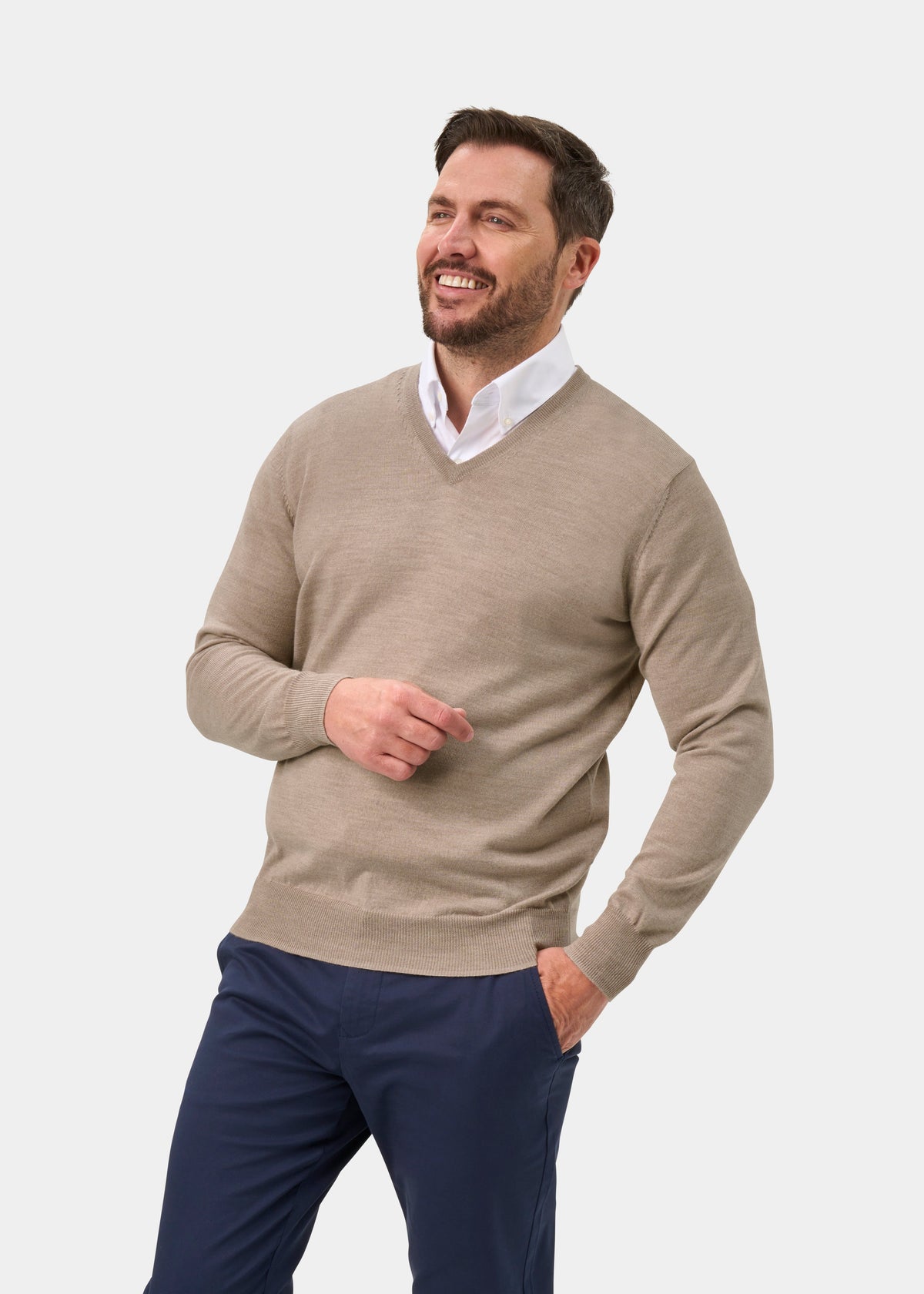 A smiling man wearing a light beige v-neck sweater over a white collared shirt and navy blue trousers, standing against a plain light gray studio background.
