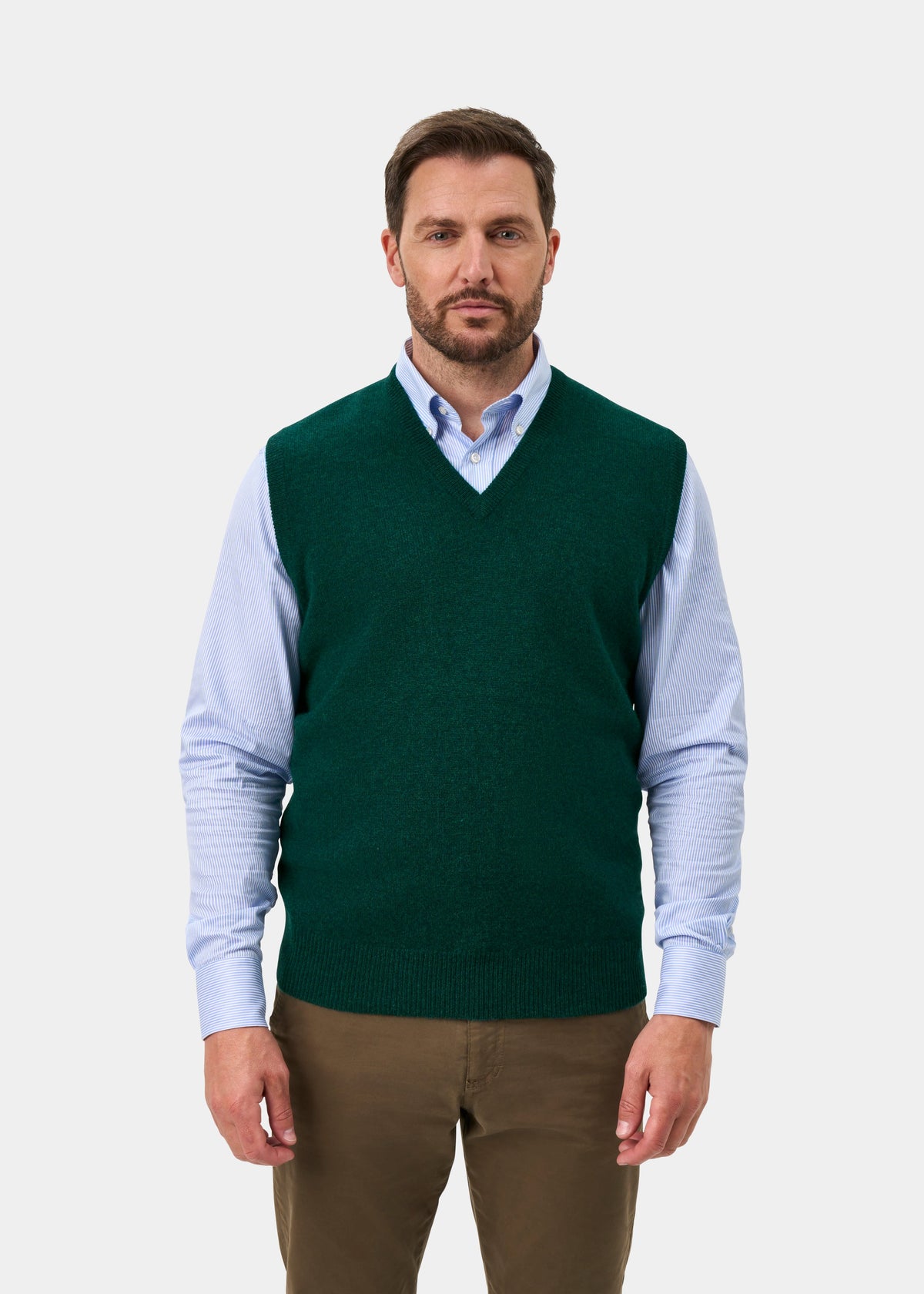 A man wearing a dark green knit sweater vest over a light blue long-sleeve button-down shirt and brown trousers, standing against a light gray studio background.