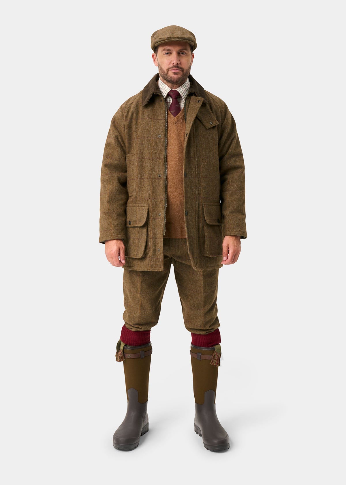 A man in a Rutland Men’s Tweed Waterproof Shooting Coat in Birch, matching breeks, flat cap, sweater, shirt, tie, and boots.
